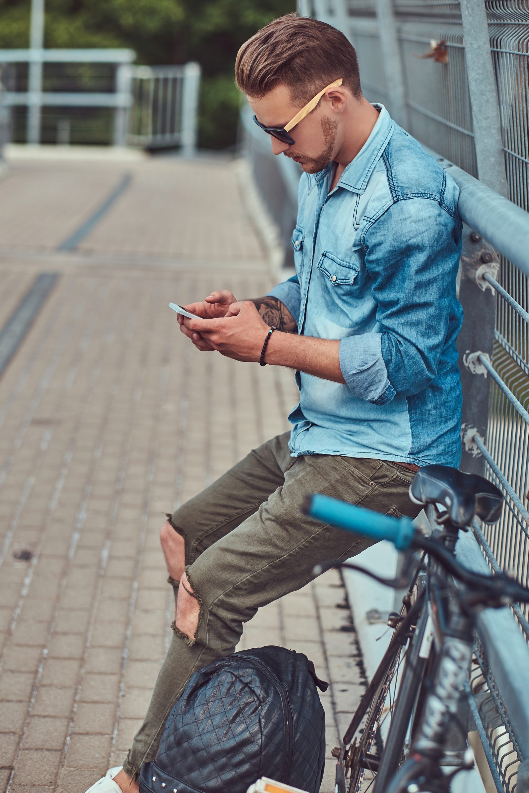 Handsome hipster with a stylish haircut in sunglasses resting after riding on a bicycle, using a smartphone.